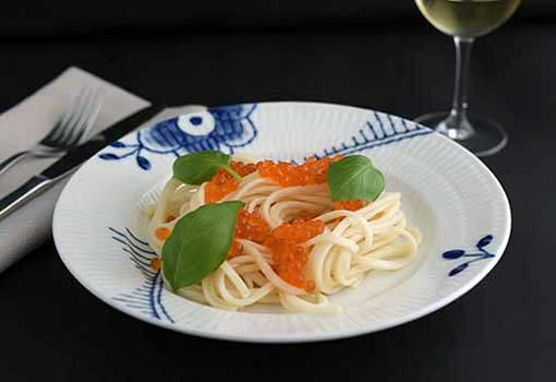 "A white plate with blue floral patterns holding a serving of linguini pasta topped with bright orange Priess & Co. trout caviar and fresh basil leaves, ready to be served." width="437" height="300"