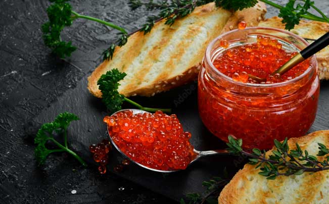 "A glass jar of vibrant red Priess & Co. Danish trout caviar, with a spoon overflowing with roe, served with golden toasted bread and fresh herbs on a dark slate background." width="651" height="404"